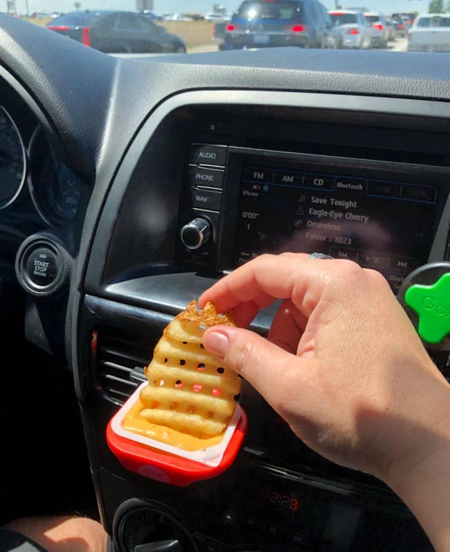 reviewer photo of them dipping a waffle fry into a packet of sauce being help by the dip clip in their car's air vent