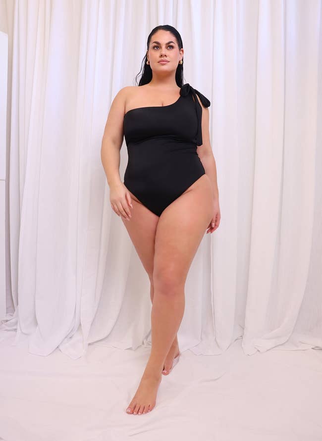 Woman in a one-shoulder black swimsuit poses against a white backdrop