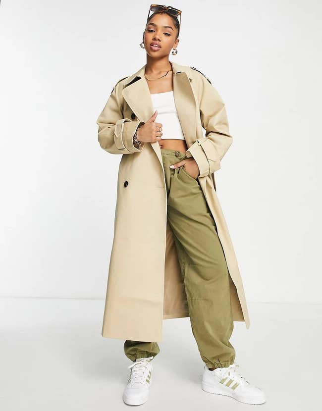 model wearing the tan coat over cargo joggers, a white crop top, and sneakers