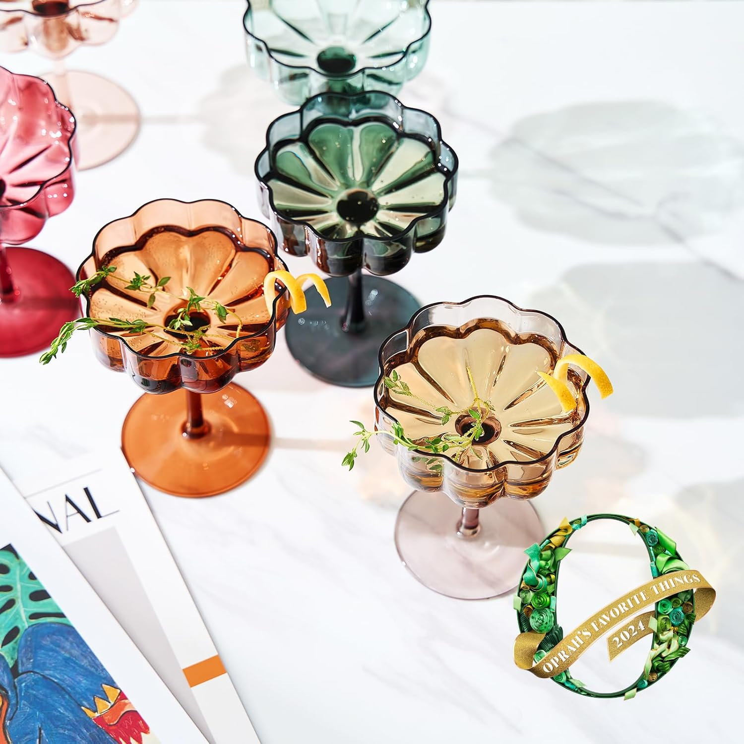 Six decorative glass cocktail cups with herbs and lemon garnishes on a table, next to a magazine and a 2023 award emblem