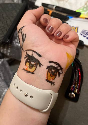 Hand with makeup test swatches drawn as eyes on the back, resting on a table with makeup items