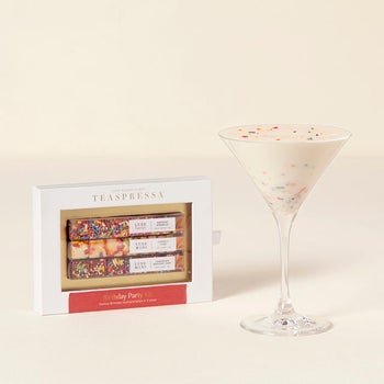 a martini glass full of a white drink with rainbow sprinkles next to the box of a birthday drink kit