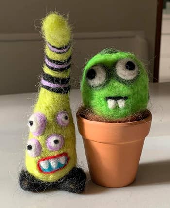 Two handmade felt monsters: one tall with stripes and different eyes, the other green with big eyes and fangs, in a small pot