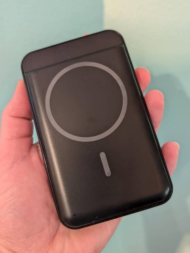 A hand holds a sleek, black portable charger with a circular design, suitable for wireless charging, against a plain background