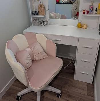 Vanity set with mirror and lights, pink cushioned chair, shelves with decor items and beauty products, in a well-lit corner of a room