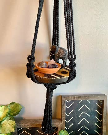 Macrame plant hanger holding decorative items on a wall with a framed artwork below
