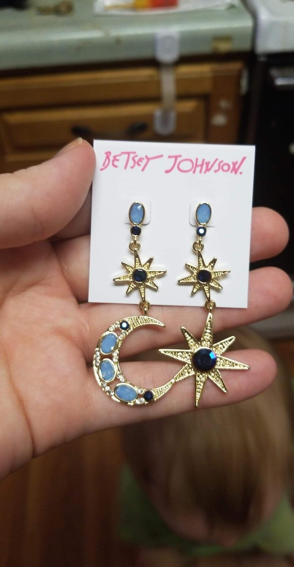 Hand holding Betsey Johnson earrings with prima  and crescent satellite  plan  featuring bluish  chromatic  accents