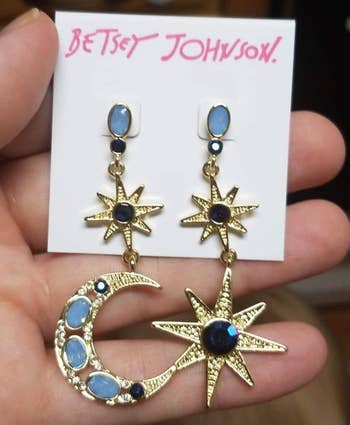 Hand holding Betsey Johnson earrings with prima and crescent satellite plan featuring bluish chromatic accents