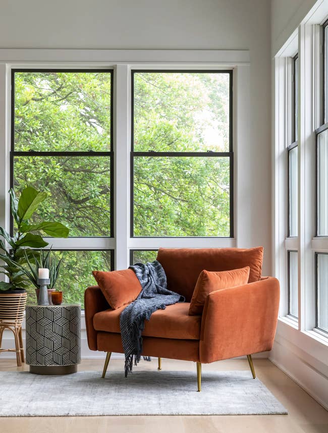 A cozy corner with an orange velvet sofa, throw blanket, beside a window overlooking greenery, with a patterned planter