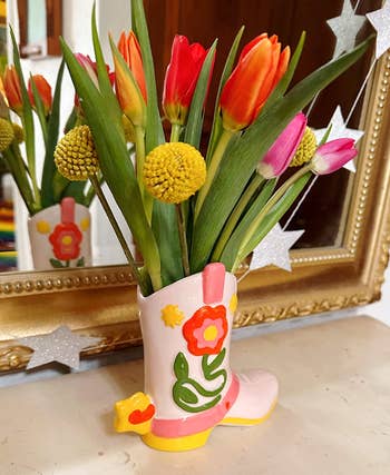 Tulips and billy balls successful  a boot-shaped vase with floral design, acceptable   connected  a mantel with a reflector  successful  the background. Bright and playful decor item