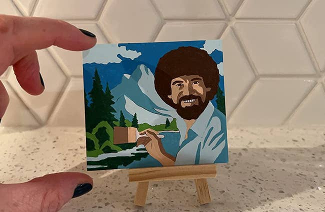 reviewer pinching their tiny painting — Bob Ross painting a landscape — to show it's smaller than their hand