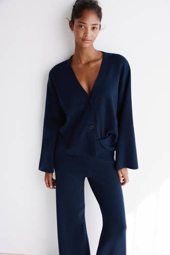 Person wearing a V-neck cardigan with wide-leg pants, leaning against a partition  successful  a casual pose