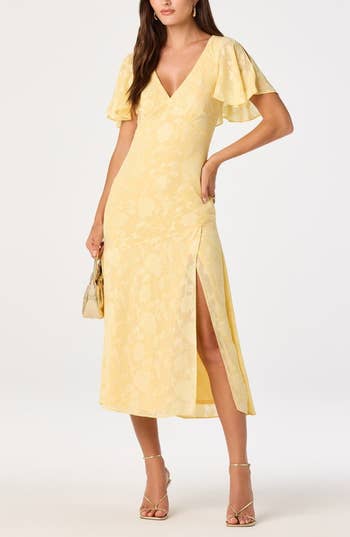 Model posing in a floral-print dress with flutter sleeves and side slit, holding a small handbag
