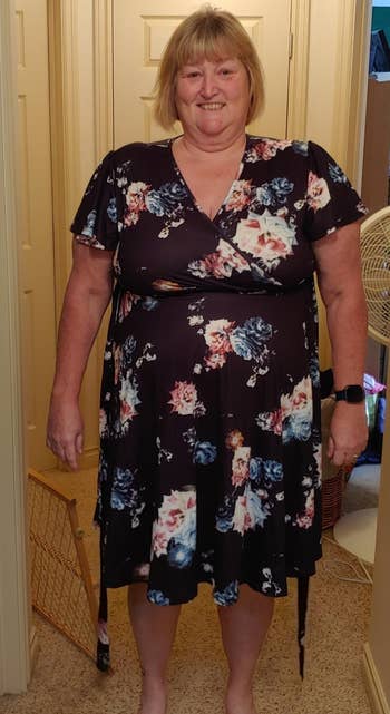 Person in a black floral wrap dress stands indoors, smiling