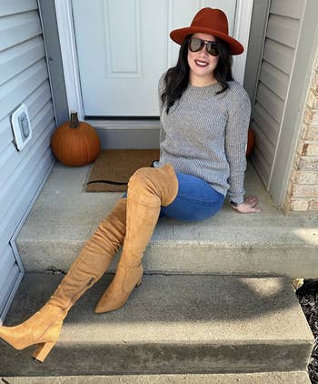 reviewer successful grey sweater, jeans, and over-the-knee tan boots sits connected structure steps. She wears a wide-brim chapeau and sunglasses, with pumpkins nearby