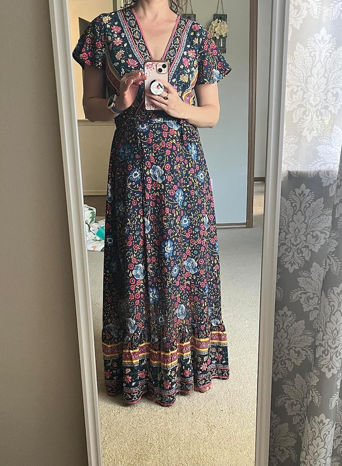 A reviewer wearing a floral maxi dress with short sleeves stands in front of a mirror while holding a phone with a decorative case