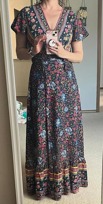 A reviewer wearing a floral maxi dress with short sleeves stands in front of a mirror while holding a phone with a decorative case