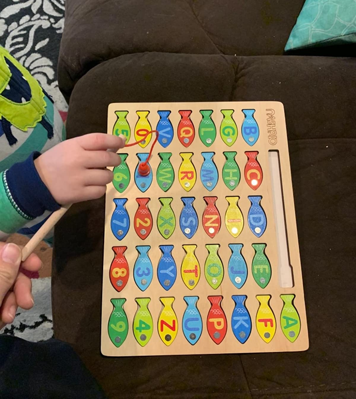 reviewer photo of a kid's hand using the magnetic fishing rod to grab a piece from the letter, number, and colored fish puzzle