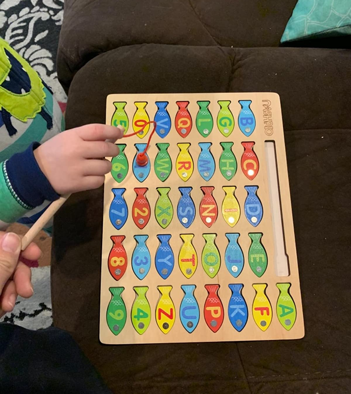 reviewer photo of a kid's hand using the magnetic fishing rod to grab a piece from the letter, number, and colored fish puzzle