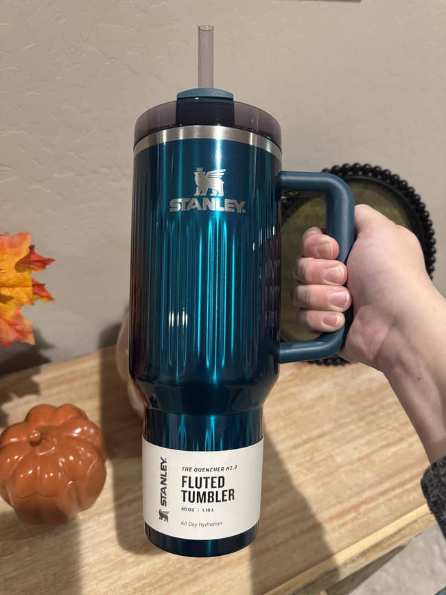 Person holding a 40 oz Stanley Quencher H2.0 Fluted Tumbler with a straw and handle; pumpkin decor successful the background