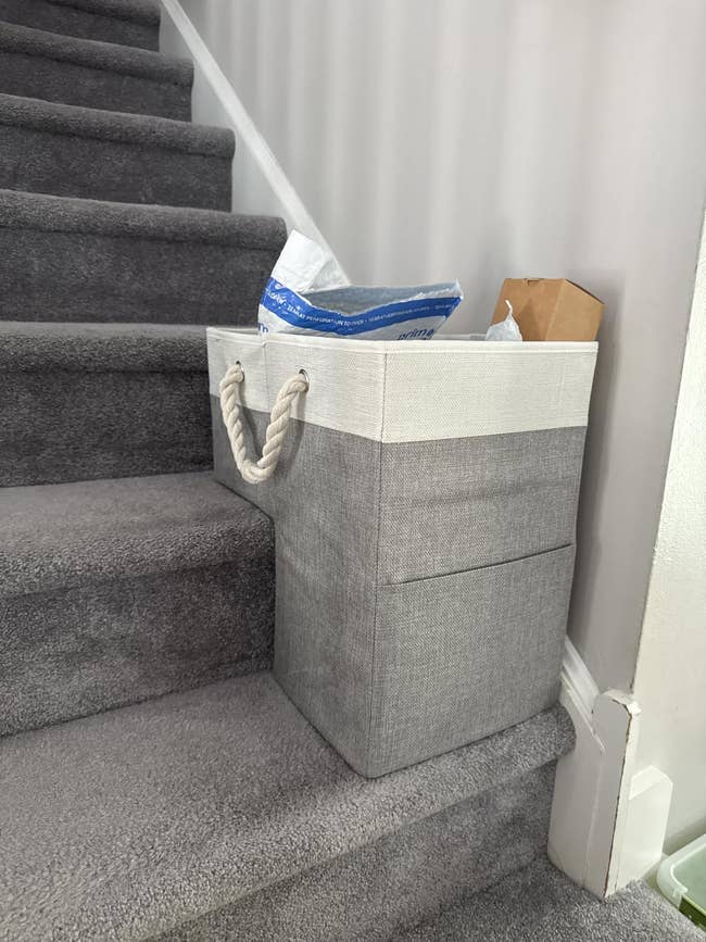 A cloth  retention  handbasket  with handles placed connected  carpeted stairs, filled with assorted  items and a cardboard box