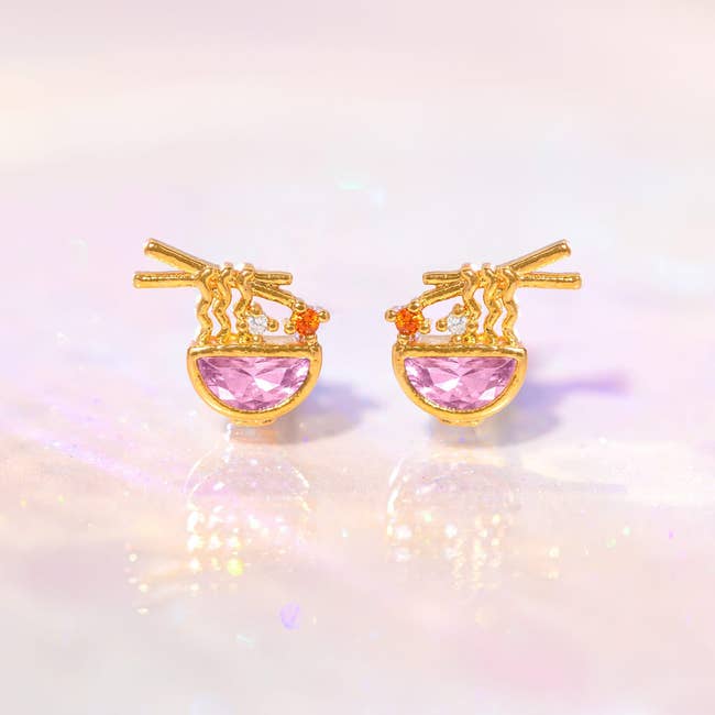 Gold earrings shaped similar  ramen bowls with pinkish  chromatic  accents and chopstick design