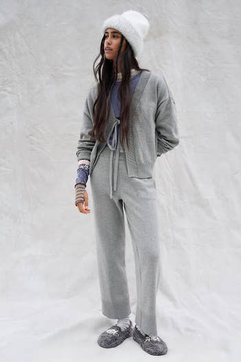 Person in cozy, casual outfit with knit cardigan, pants, hat, and slippers stands against a plain backdrop, featured in shopping article