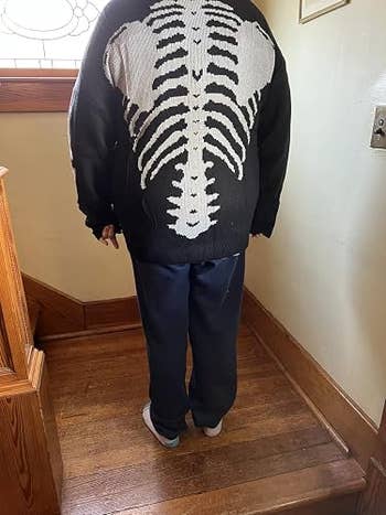 Person stands indoors wearing a sweater with a skeleton plan  connected  the backmost  and acheronian  pants, suitable for casual, cozy buying  attire