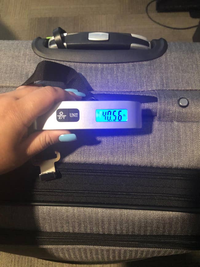 A hand holds a portable scale showing the weight of a suitcase as 40.56 pounds