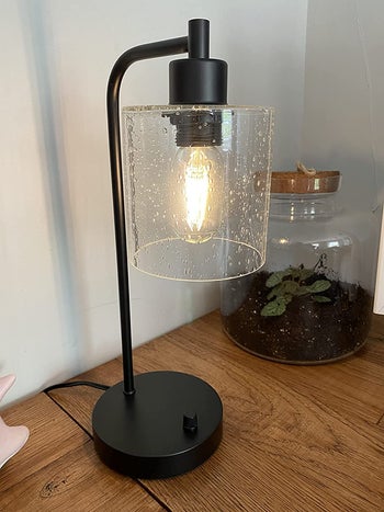 reviewer photo of a black industrial-style desk lamp with a glass shade and the bulb turned on