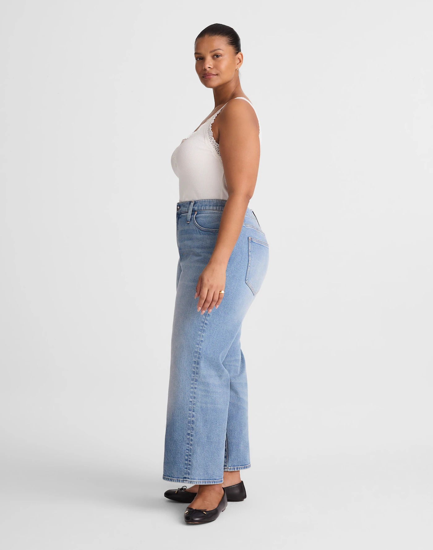 A person poses sideways wearing high-waisted jeans and a sleeveless top, with black ballet flats, against a plain background