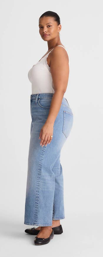 A person poses sideways wearing high-waisted jeans and a sleeveless top, with black ballet flats, against a plain background
