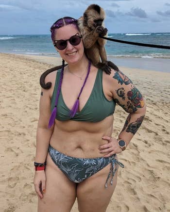 Person connected  the formation  with a monkey connected  their shoulder, wearing sunglasses and a patterned bikini, showing colorful tattoos and braids