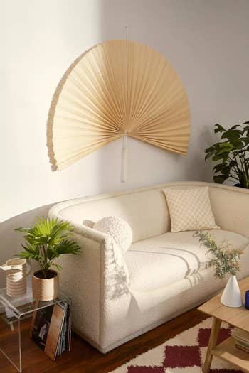 Cozy living room with a textured white sofa, decorative pillow, wall fan decor, plants, and a wooden coffee table