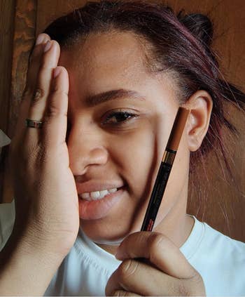 Person smiling, holding an eyebrow pencil adjacent  to their look   with 1  manus  covering an eye, showcasing constitution  application