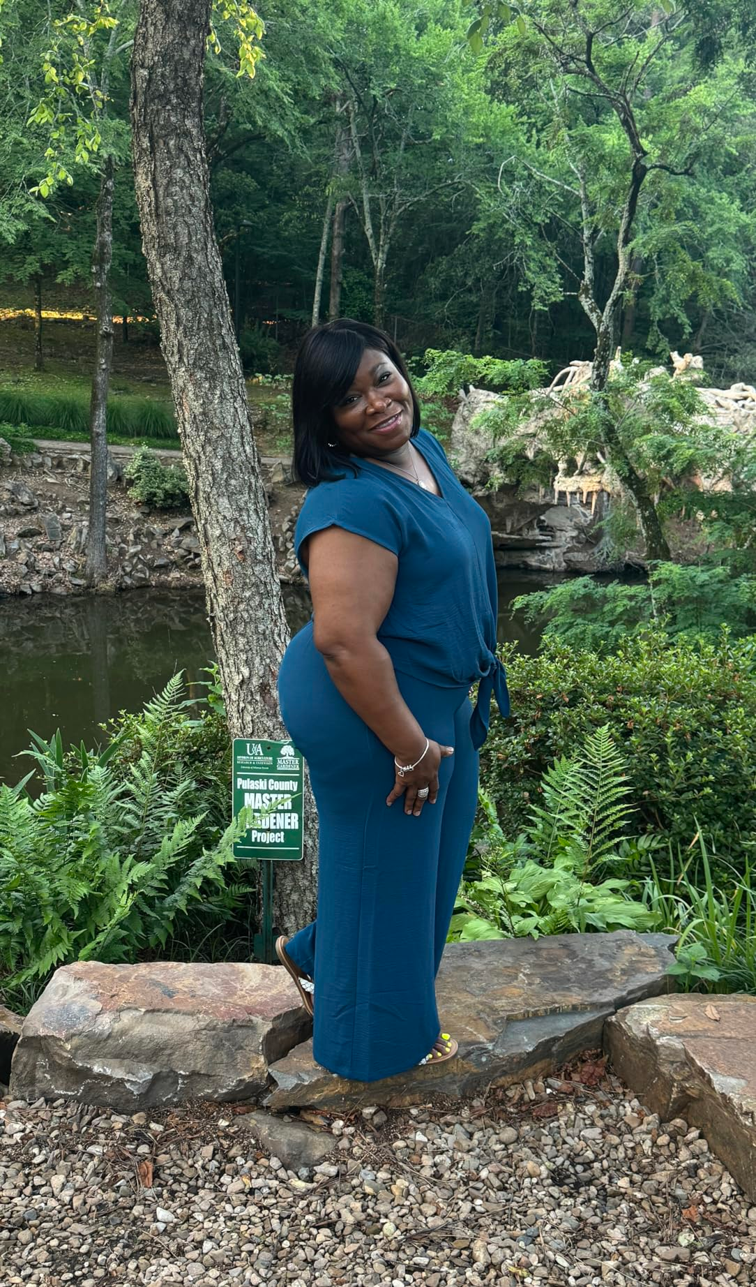Person successful  a stylish, flowy outfit smiles outdoors, lasting  connected  a rock-lined way  with lush greenery and a pond successful  the background