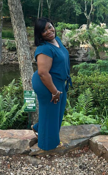 Person successful a stylish, flowy outfit smiles outdoors, lasting connected a rock-lined way with lush greenery and a pond successful the background