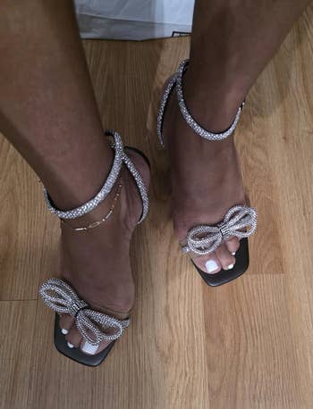 Close-up of feet wearing square-toed sandals adorned with rhinestone bow designs astir the ankles and connected the toes, connected a woody floor