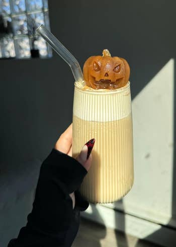 Person holding a glass with iced coffee, topped with a small pumpkin decoration and a clear straw.
