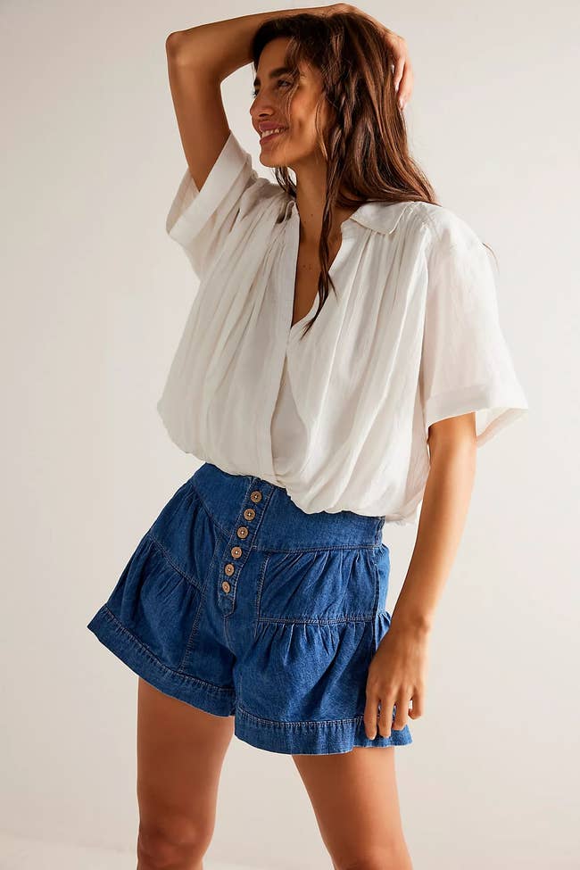 Model in a loose, white blouse and denim shorts poses with one arm raised, showcasing casual chic summer fashion