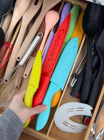 A hand opens a kitchen drawer revealing various utensils including colorful Cuisinart knives