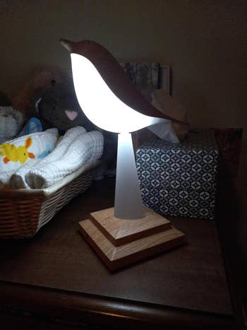 reviewer's bird-shaped lamp with illuminated body on a wooden base, placed on a shelf near a basket and plush toys