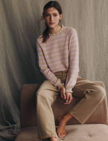 Woman successful  striped sweater and khaki pants sits connected  a sofa, looking astatine  the camera