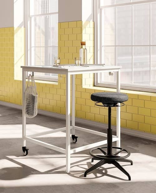 Tall table with wheels, holding a bottle and glasses, stands in a sunlit room. Adjacent stool with round seat included. Bright setting