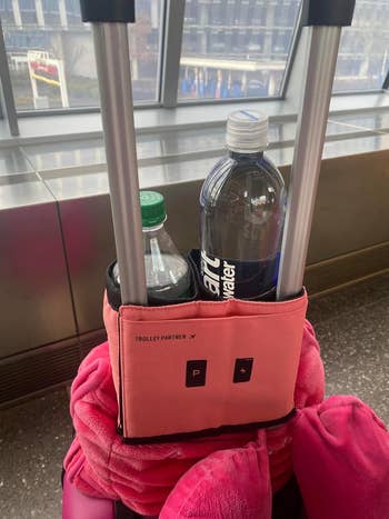 A travel trolley with a coral organizer holding two water bottles and a pink scarf, by a window at an airport