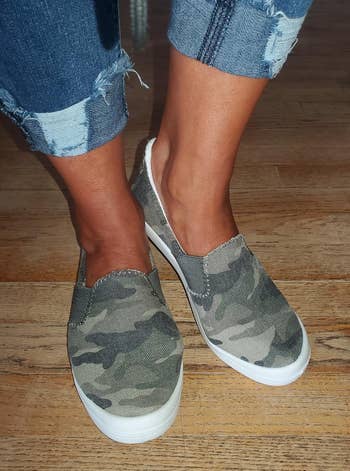 a reviewer wearing slip-on sneakers featuring a camouflage print 
