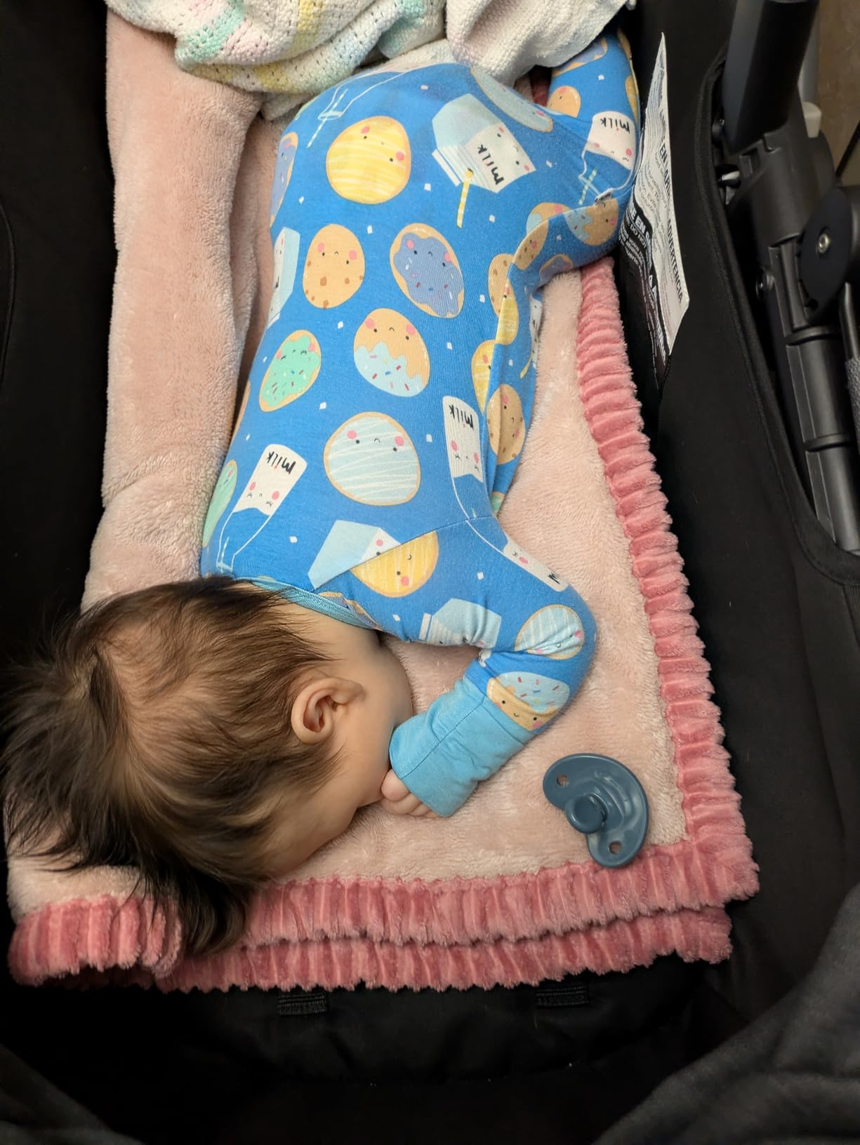 Baby sleeping successful  a stroller connected  a pinkish  blanket, wearing a patterned onesie with planets. A pacifier lies nearby