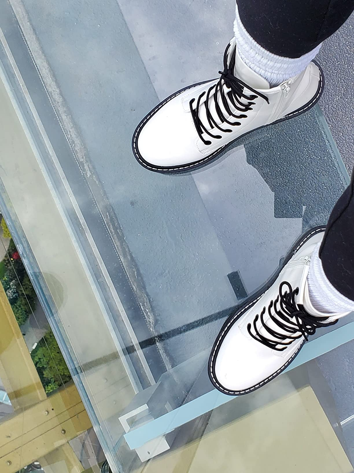 Person wearing white lace-up boots standing on a clear glass floor, offering a view of the ground below