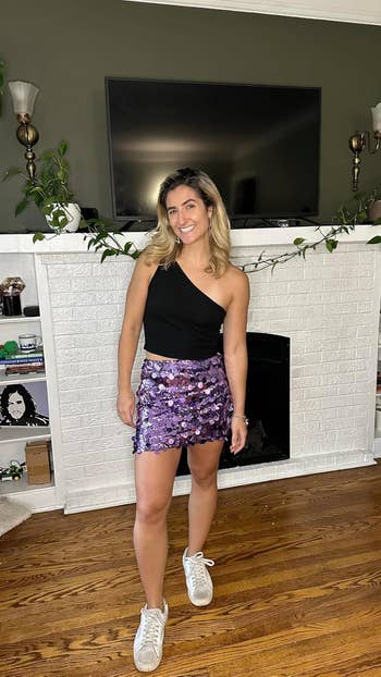 Reviewer in a black one-shoulder top, purple sequined miniskirt, and white sneakers poses in front of a white brick fireplace with plants