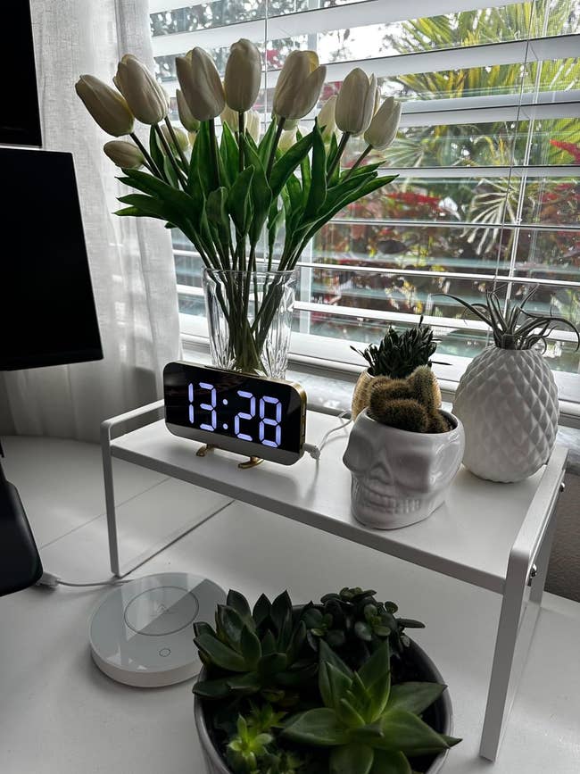 A modern desk with a digital clock, a bouquet of tulips, a small potted plant, and decorative items